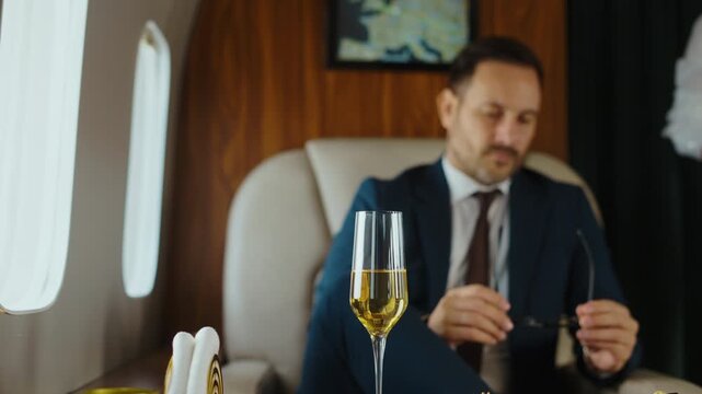 Confident wealthy man in a suit relaxing on a luxury private airplane, putting on glasses and toasting with a champagne glass while enjoying a delicious meal during a first-class flight