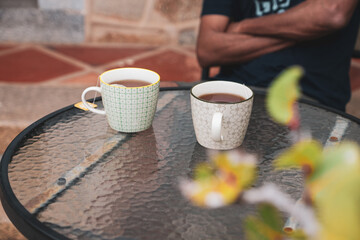 Close-up of two ceramic cups with morning tea on glass table with cropped image of a South Asian man sitting hand-crossed. Selective focus on the cup.