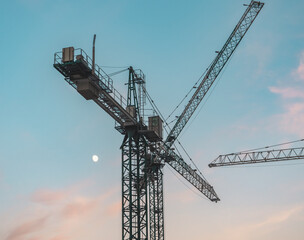 Crane silhouettes against pastel twilight sky with moon over construction site during evening