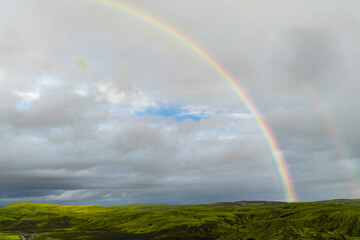 Naklejka premium A bright rainbow arcs above moss green lava fields near Blue Lagoon in southwest Iceland. Dark basalt patches and a winding stream sit under clouds breaking to blue.