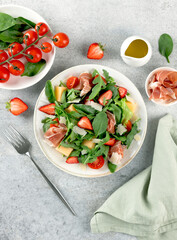 Italian arugula salad with prosciutto, strawberries, melon, cheese on gray background. Top view. 