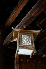 A detailed shot of an old, ornate wooden lantern hanging from the dark beams of a Japanese temple or shrine, featuring a carved chrysanthemum crest on its panel.