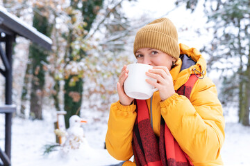 Happy woman in yellow jacket and hat drinks hot beverage from white mug outdoors on snowy day, enjoying warmth and peace in a winter landscape. Winter relaxation and mindful living