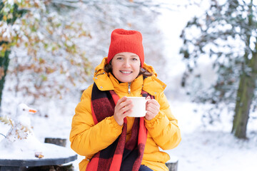 Smiling woman in red hat and yellow jacket enjoys hot drink from white mug in snowy park, radiating warmth and joy during cold winter. Self-care and happiness in outdoor leisure. Holiday warmth