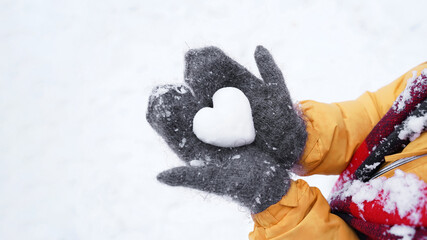 Human hands in warm gray crocheted mittens with snowy heart against snow background, vulnerability of love, fragility and transience of emotions. St.Valentine's Day concept. Warmth in cold weather