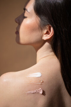 Brunette woman with long dark hair poses with skincare products applied on her shoulder, showcasing different textures and colors, highlighting beauty and self-care concepts