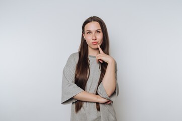 A young woman with long hair, wearing a gray t-shirt, looking thoughtful with her finger on her chin against a plain background