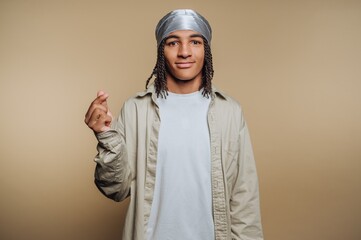 A young person with braided hair wearing a silver headscarf and a light-colored shirt, making a gesture with their hand against a neutral background