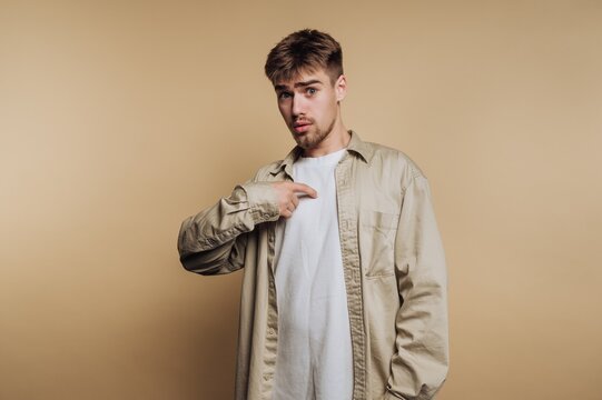 Young man in casual clothing pointing at himself against a beige background.