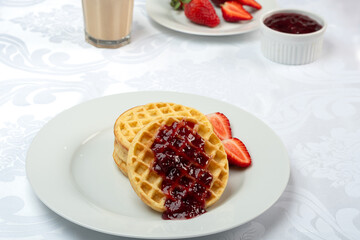 Delicious waffles with strawberry jam and fresh red berries. A glass of coffee with milk in the background.