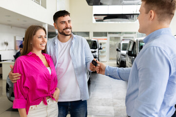 Obraz premium Smiling car salesman handing key to cheerful couple in dealership. Happy buyers receiving vehicle keys after successful purchase in bright auto showroom