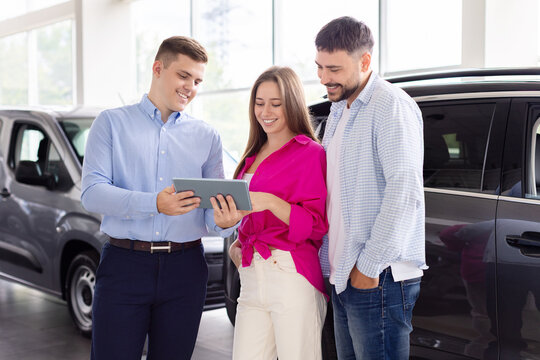 Salesman showing details on digital tablet to smiling couple in a car dealership. Friendly communication and technology-driven customer experience in showroom - Powered by Adobe