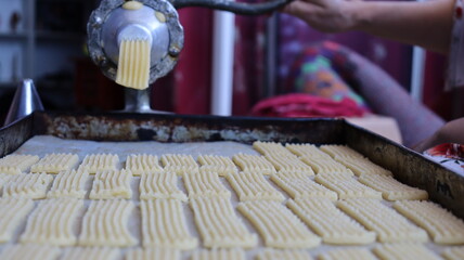 Halwa Lbouq (Moroccan Horn Pastry) - Batch of Sesame-Coated Pastries Ready to Bake, Woman Shaping Raw Dough with Traditional Cookie Press, Silver Bracelets, Home Baking Setup for Eid/Ramadan