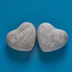 Two heart-shaped stones lie side by side on a solid blue background. One reads  BE HAPPY and the...