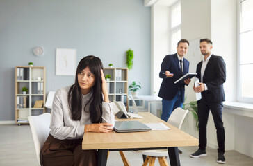A woman sits at a desk in an office, showing stress and loneliness as colleagues chat, highlighting isolation in the workplace and a business conflict. Quiet tension affects focus and morale.