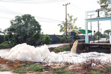 Murky foam piles created by floating fast from flow of polluted water from a large water pump to accelerate drainage after rain at irrigation canal gate along village road in rural Thailand.