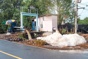 Murky foam piles created by floating fast from flow of polluted water from a large water pump to accelerate drainage after rain at irrigation canal gate along village road in rural Thailand.