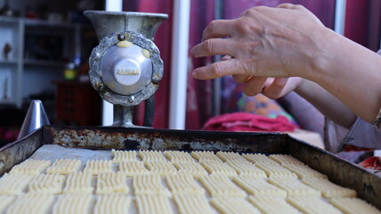Halwa Lbouq (Moroccan Horn Pastry) - Batch of Sesame-Coated Pastries Ready to Bake, Woman Shaping Raw Dough with Traditional Cookie Press, Silver Bracelets, Home Baking Setup for Eid/Ramadan