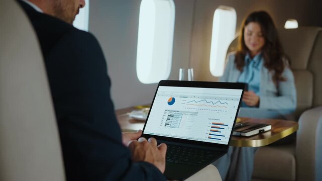 Focused businessman analyzing financial data on a laptop while traveling on a luxurious private jet, with a female colleague sitting across the table using her smartphone during the business trip