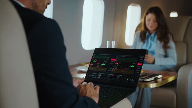 Focused businessman working on a laptop, checking stock market charts and financial data while traveling on a luxurious private jet with a female colleague sitting in front of him