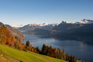 A spectacular Alpine vista over a deep blue lake, framed by sunlit green slopes and vivid autumn forests, with snow-capped mountains and a clear sky.