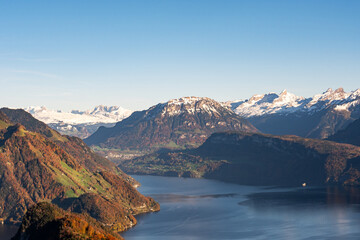 A spectacular mountain panorama showcases a winding lake nestled among rugged, snow-dusted autumn slopes under a clear blue sky.