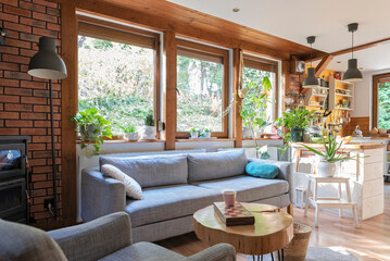 Rural cottage with cozy design, windows to the forest, fireplace, sofa, armchair and red brick wall. Interior of living room with design indoor architecture.
