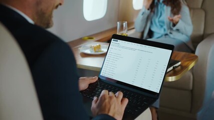 Successful businessman traveling in a luxurious private jet, analyzing financial charts and stock market data on his laptop while a colleague talks on the phone in the background - Powered by Adobe