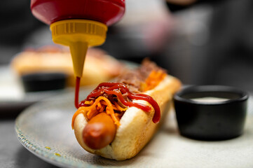 A delicious hot dog is being topped with ketchup and other condiments on a plate, with a focus on...