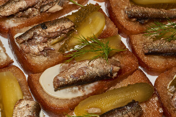 Close-up of delicious sandwiches with canned sprats, pickled cucumbers, and fresh herbs on toasted rye bread. Classic retro-style snack: sprats on crispy bread with mayonnaise and pickled cucumber.