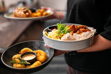 A person holds two bowls of delicious fried food, including chicken wings, calamari, fried shrimp, and fries, with dipping sauces and fresh herbs
