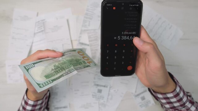 Hands holding cash and a smartphone with a calculator app, with numerous receipts scattered on a desk. This image represents personal finance management and digital money tracking