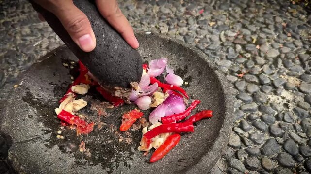 The making of chili paste called sambal , grinding red chili peppers, onion, and garlic in a rough stone mortar and pestle on a cobblestone surface, conveying rustic cooking and fresh spices