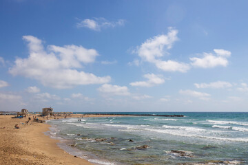 Haifa, Israel, A  beach on a clear day, with moderate waves breaking on the shore near a small rock formation, people enjoying the sand, and a small complex of buildings with thatched umbrellas