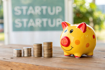A bright yellow piggy bank sits next to growing stacks of coins on a wooden surface, against a blurred backdrop of the word "STARTUP," symbolizing initial business funding