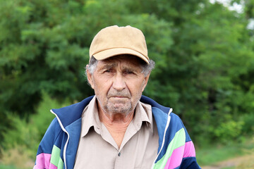 Elderly man looks into camera on forest background. Sad face expression, concept of life in village, old age and retirement
