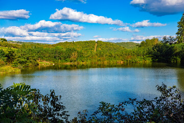 Fototapeta premium 2025-10-02 CALM LAKE FRAMED AND SURROUNDED BY GREEN FOLIAGE AND A ROLLING HILL IN THE BACKGROUND AND A NICE CLOUDY SKY NEAR SIENA IN THE TUSCUNY REGION OF ITALY