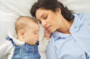 Close portrait of a mother gazing at her sleeping infant on a white bed, showing maternal love and family connection in soft light. Mother watches sleeping baby with gentle affection.