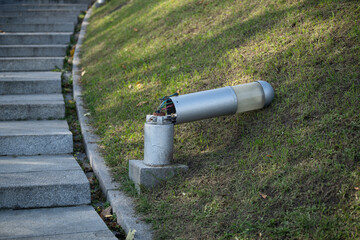 A broken electric lantern stands next to a set of stone steps in a sunny city park. The device...