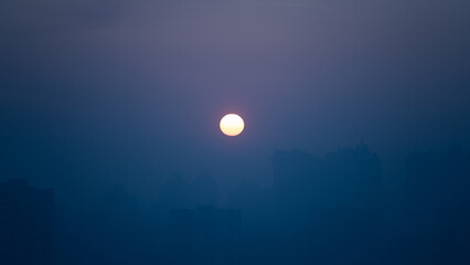 Obraz premium A warm sun sets in the evening, surrounded by a hazy sky that provides a tranquil backdrop. The city skyline is barely visible, enhancing the peaceful scene