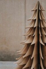 A minimal Christmas tree crafted from recycled cardboard panels forms a stylized layered structure standing against a plain neutral wall in a simple and eco-friendly design
