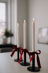A trio of tall white taper candles placed in matte black holders each decorated with red velvet bows, arranged on a neutral table in a modern Christmas interior
