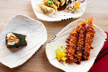 Japanese traditional snacks, prawn tempura, onigiri and gyoza served on white plates.