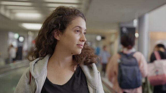 Curly-haired woman sips coffee in airport terminal. Soft focus background shows travelers moving. Tired but happy. Traveling abroad for vacation. Standing on moving walkway on way to departure gate