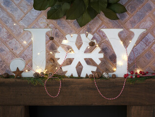 Brick fireplace hearth mantle Christmas holiday festive decoration with white JOY block farmhouse word letters and fairy lights, green wreath and brown wood shelf.