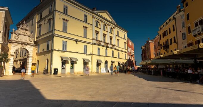 Rovinj, Croatia - August 15, 2025: Timelapse, slide transition. Sunlit square in Rovinj with pastel-colored buildings, a stone archway, and a lively caf&eacute; terrace filled with people.