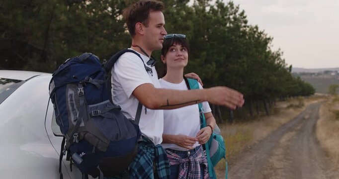 Smiling hiking couple by car with backpacks. They pause on forest road, enjoying outdoor travel before journey on pine trail, hikers. Concept freedom, outdoor adventure and road trip.