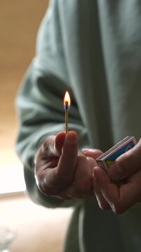 A smoldering match in the hands of a man in a blue sweater.
A man in the kitchen lights a wooden match against a matchbox.