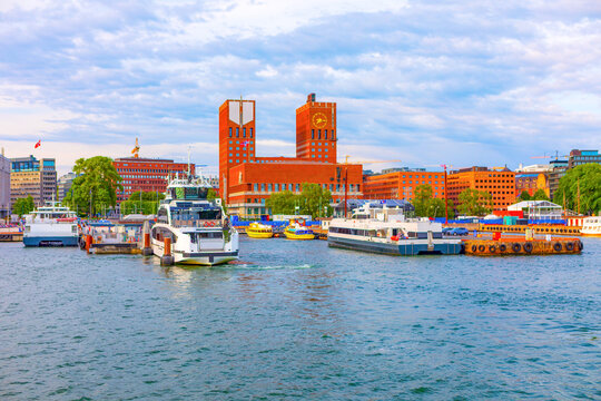 Oslo City Hall prominent brick building with two towers, overlooking the harbor in Oslo, Norway, boats docked in foreground. Cloudy sky casting soft light on city waterfront and surrounding buildings