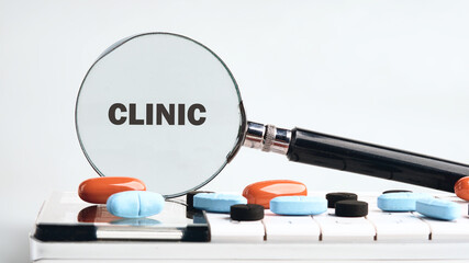 The magnifying glass is focusing on the word Clinic. A keyboard sits below the pills of different colors. It is a bright concept that alludes to healthcare services